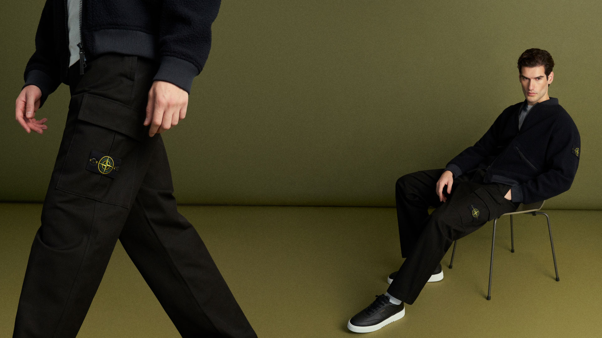 Mann sitzt auf Stuhl, trägt schwarze Cargo-Hose mit Logo und dunkle Jacke, weitere Person läuft im Vordergrund vorbei. Mann sitzt auf Stuhl, trägt schwarze Cargo-Hose mit Logo und dunkle Jacke, weitere Person läuft im Vordergrund vorbei.