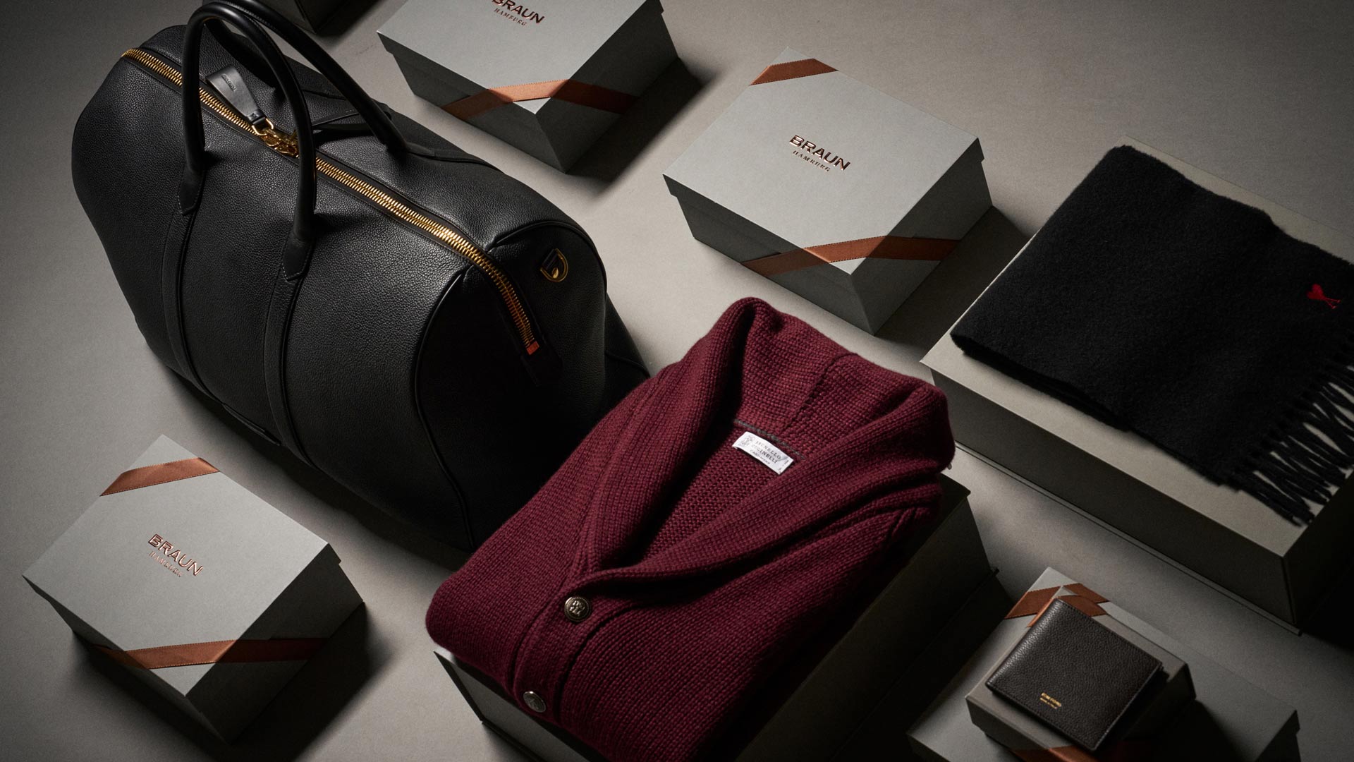 Schwarze Lederreisetasche, bordeauxroter Strickpullover, schwarzer Schal und mehrere graue Geschenkboxen mit Braun-Logo. Schwarze Lederreisetasche, bordeauxroter Strickpullover, schwarzer Schal und mehrere graue Geschenkboxen mit Braun-Logo.