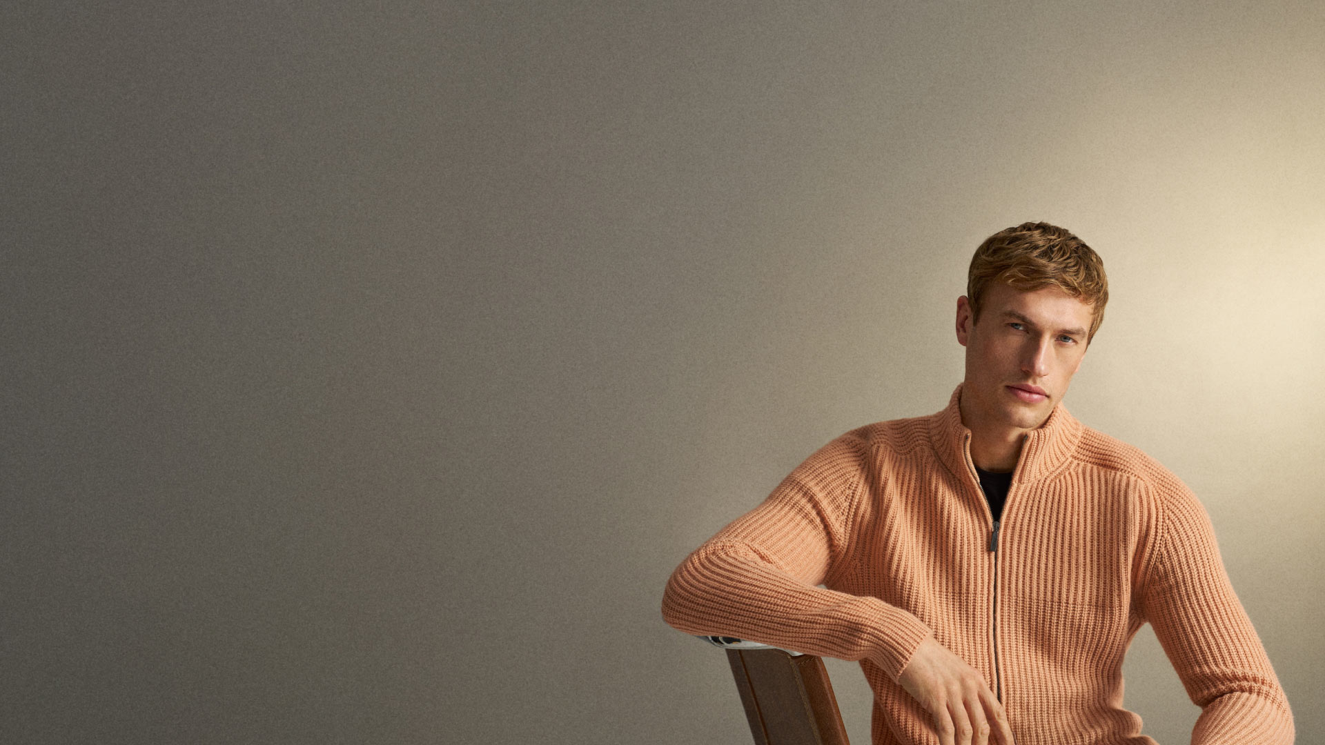 Junger Mann mit kurzen blonden Haaren trägt einen hellorangen Strickpullover und sitzt entspannt auf einem Stuhl vor grauem Hintergrund. Junger Mann mit kurzen blonden Haaren trägt einen hellorangen Strickpullover und sitzt entspannt auf einem Stuhl vor grauem Hintergrund.