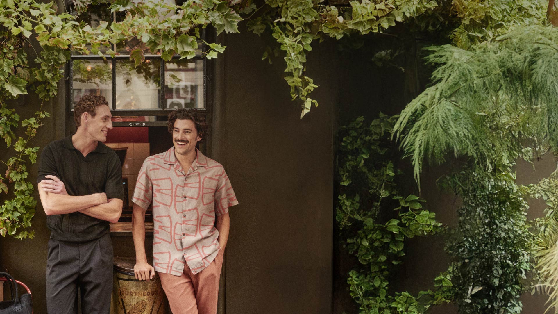 Zwei Männer unter grünen Blättern lehnen entspannt an einer Wand neben einem Fenster, sie unterhalten sich lachend. Zwei Männer unter grünen Blättern lehnen entspannt an einer Wand neben einem Fenster, sie unterhalten sich lachend.