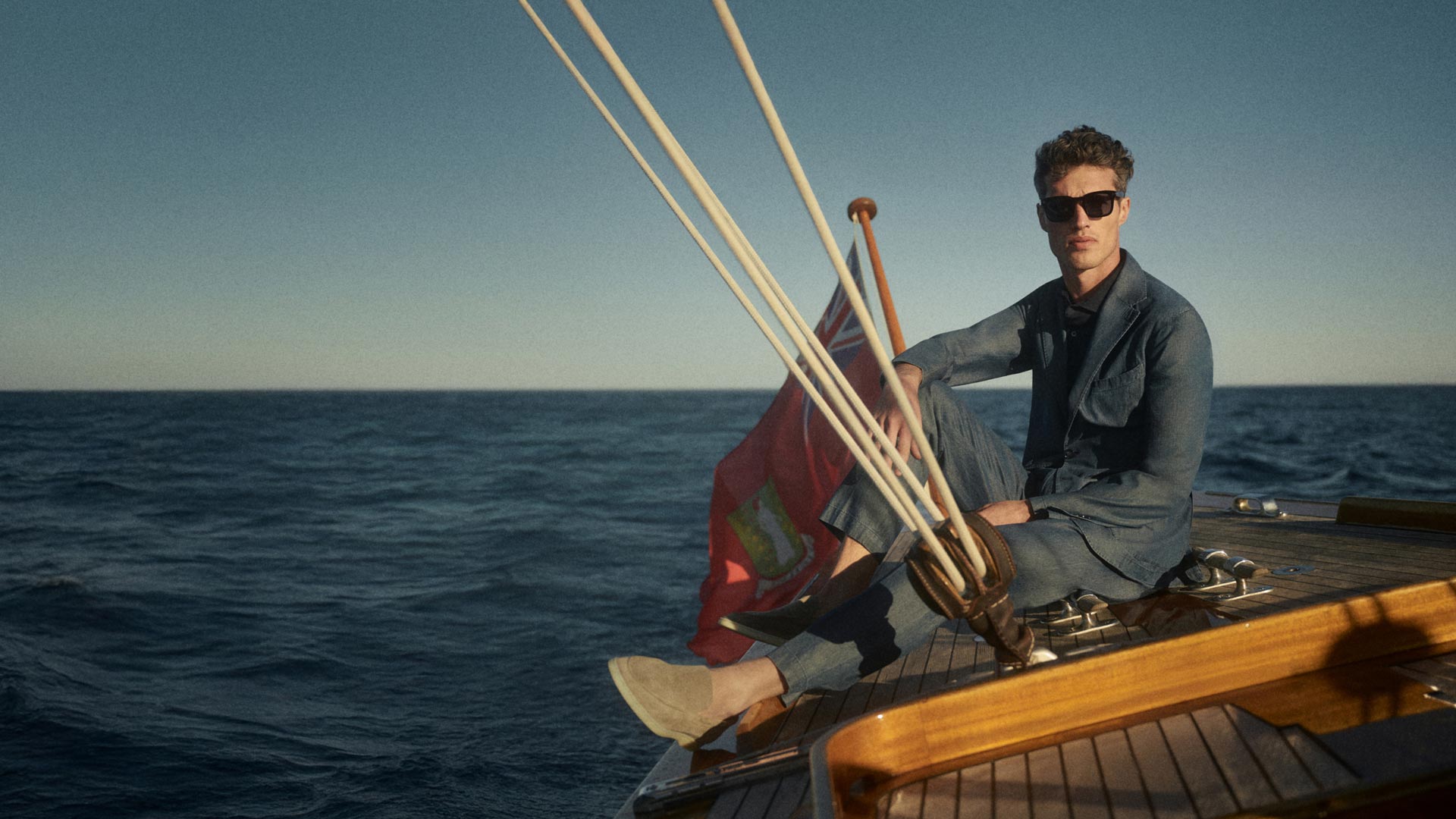 Mann mit Sonnenbrille in blauem Anzug sitzt auf Segelboot-Deck, ruhiges Meer und klarer Himmel im Hintergrund. Mann mit Sonnenbrille in blauem Anzug sitzt auf Segelboot-Deck, ruhiges Meer und klarer Himmel im Hintergrund.