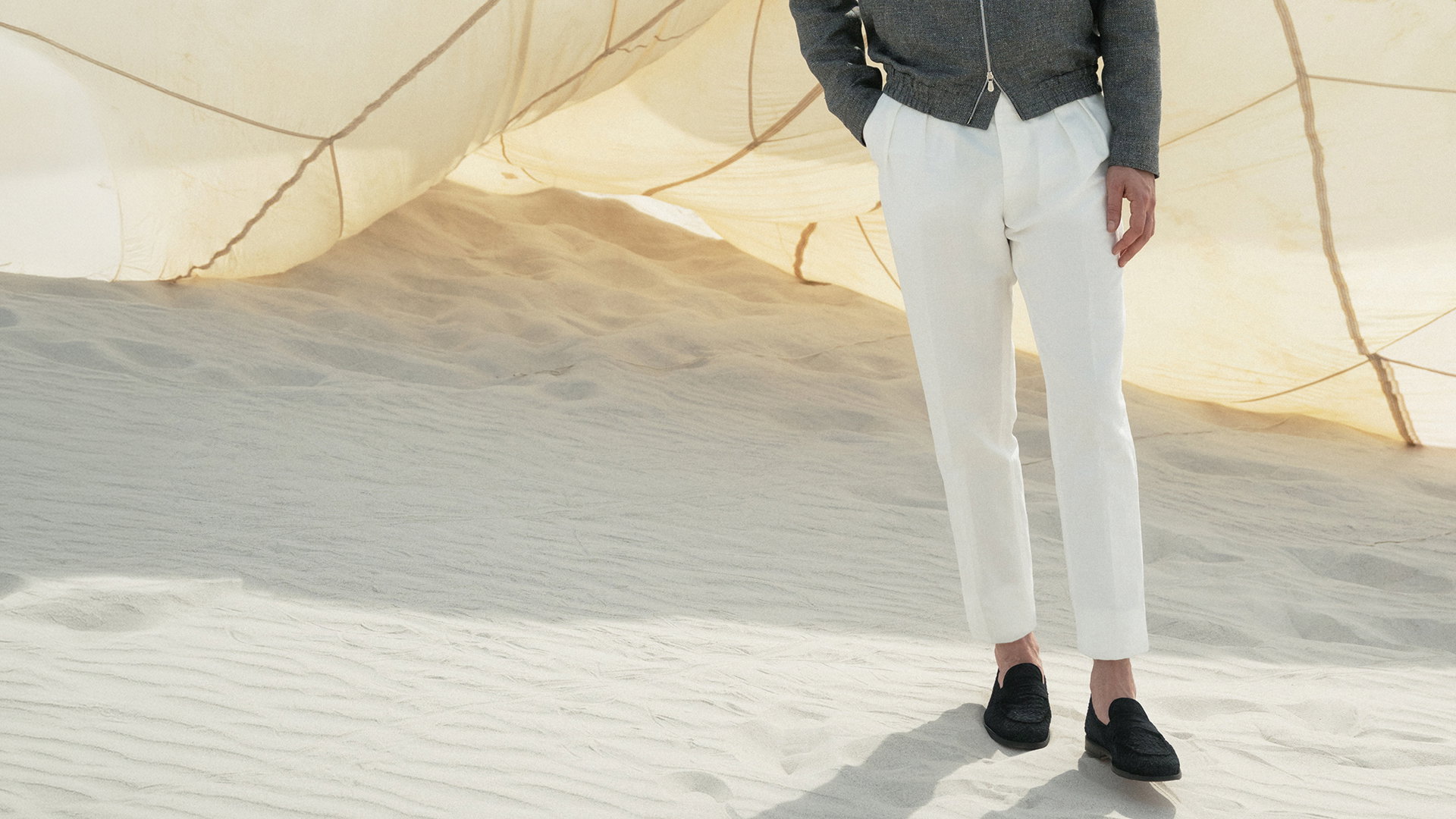 Mann trägt weiße Hose, schwarze Schuhe und graue Jacke, steht auf hellem Sand vor einem großen, hellen Segeltuch. Mann trägt weiße Hose, schwarze Schuhe und graue Jacke, steht auf hellem Sand vor einem großen, hellen Segeltuch.