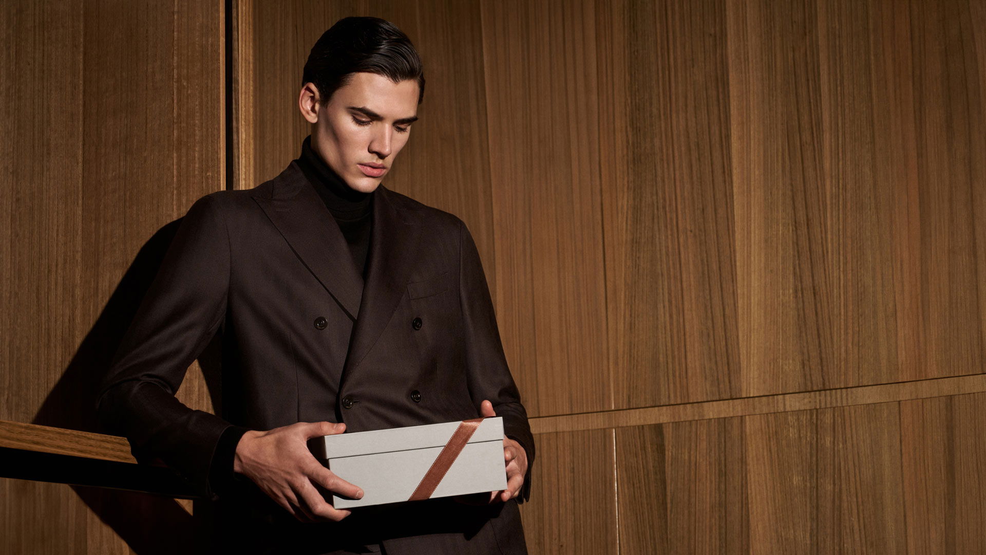 Junger Mann in dunkelbraunem Anzug hält Geschenkbox mit braunem Band vor holzvertäfelter Wand. Junger Mann in dunkelbraunem Anzug hält Geschenkbox mit braunem Band vor holzvertäfelter Wand.