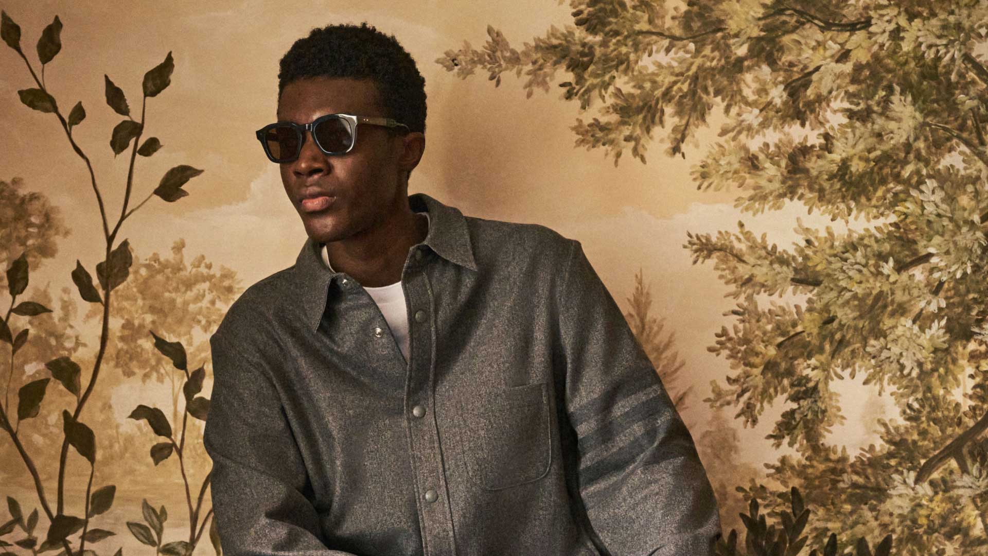 Junger Mann mit dunkler Hautfarbe trägt graues Hemd und Sonnenbrille, vor gemaltem Hintergrund mit Blättern und Bäumen. Junger Mann mit dunkler Hautfarbe trägt graues Hemd und Sonnenbrille, vor gemaltem Hintergrund mit Blättern und Bäumen.