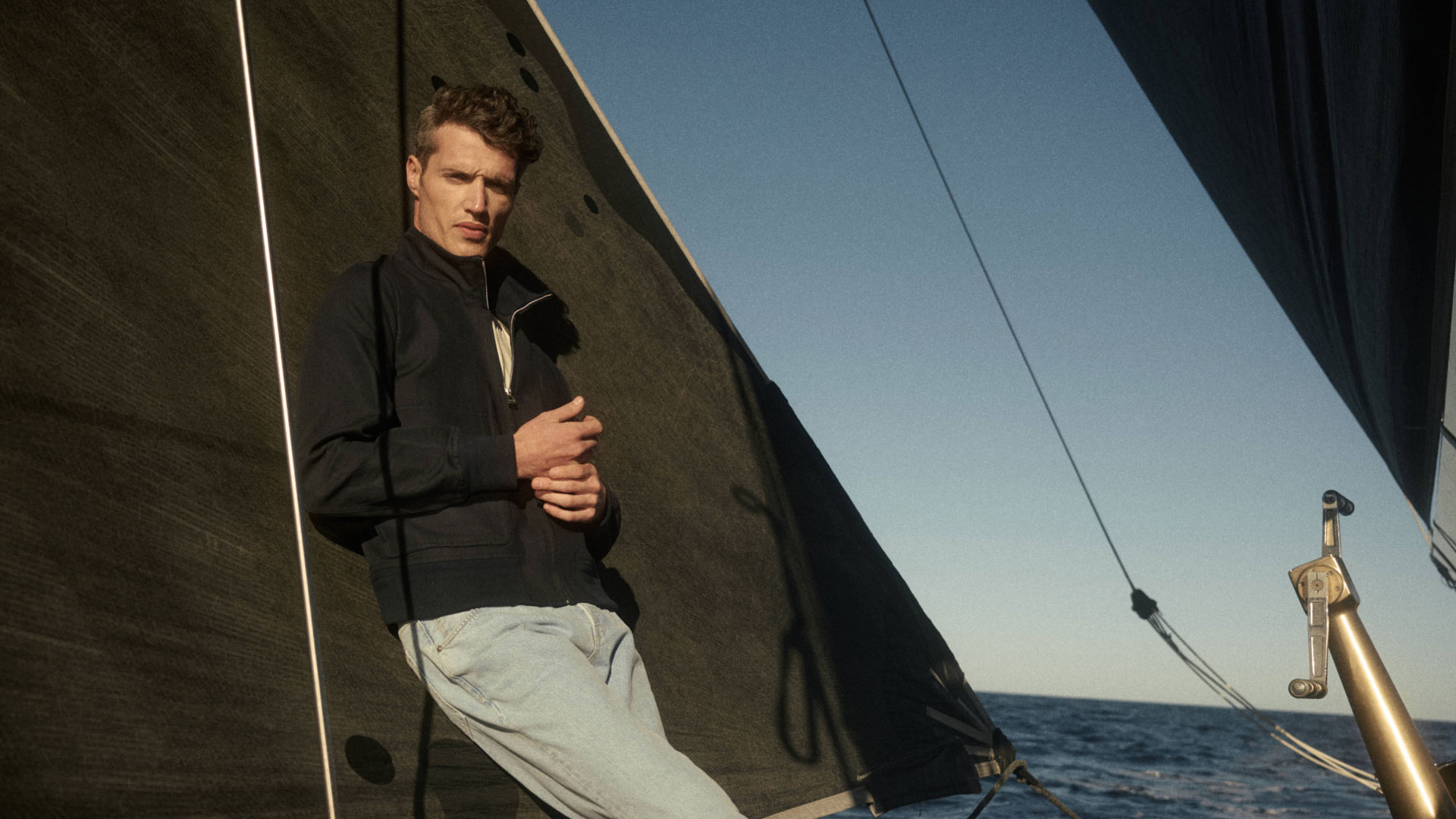 Man in black jacket and light jeans leaning against a dark sail on a sailboat with ocean and clear sky in the background. Man in black jacket and light jeans leaning against a dark sail on a sailboat with ocean and clear sky in the background.