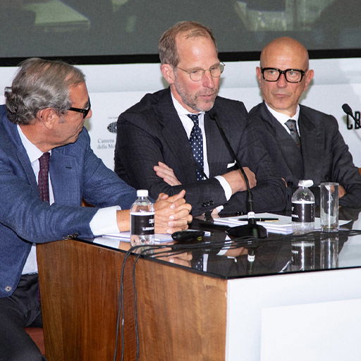 Drei Männer in Anzügen sitzen an einem Tisch mit Mikrofonen und Wasserflaschen bei einer Podiumsdiskussion. Drei Männer in Anzügen sitzen an einem Tisch mit Mikrofonen und Wasserflaschen bei einer Podiumsdiskussion.