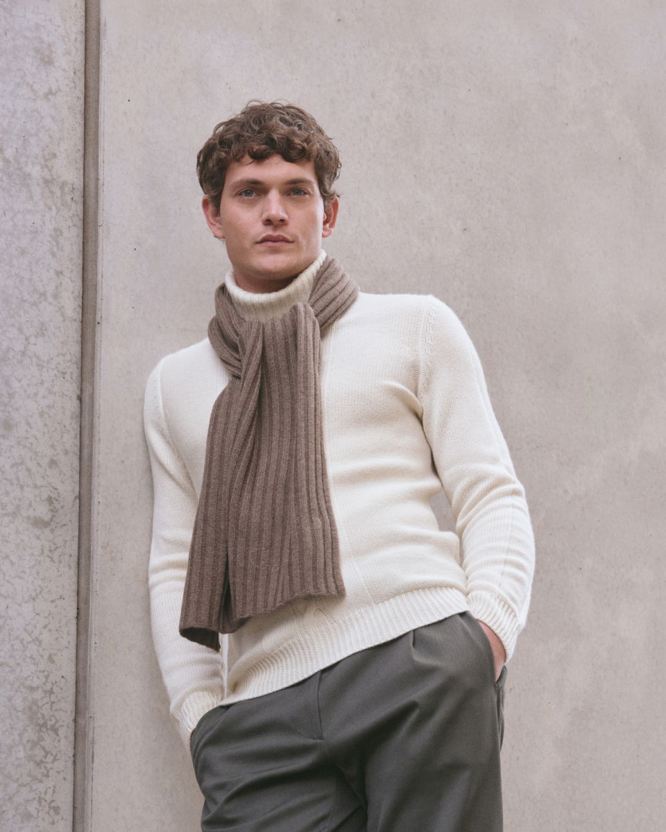 Young man wearing a cream turtleneck sweater and a brown ribbed scarf, posing against a light gray concrete wall.