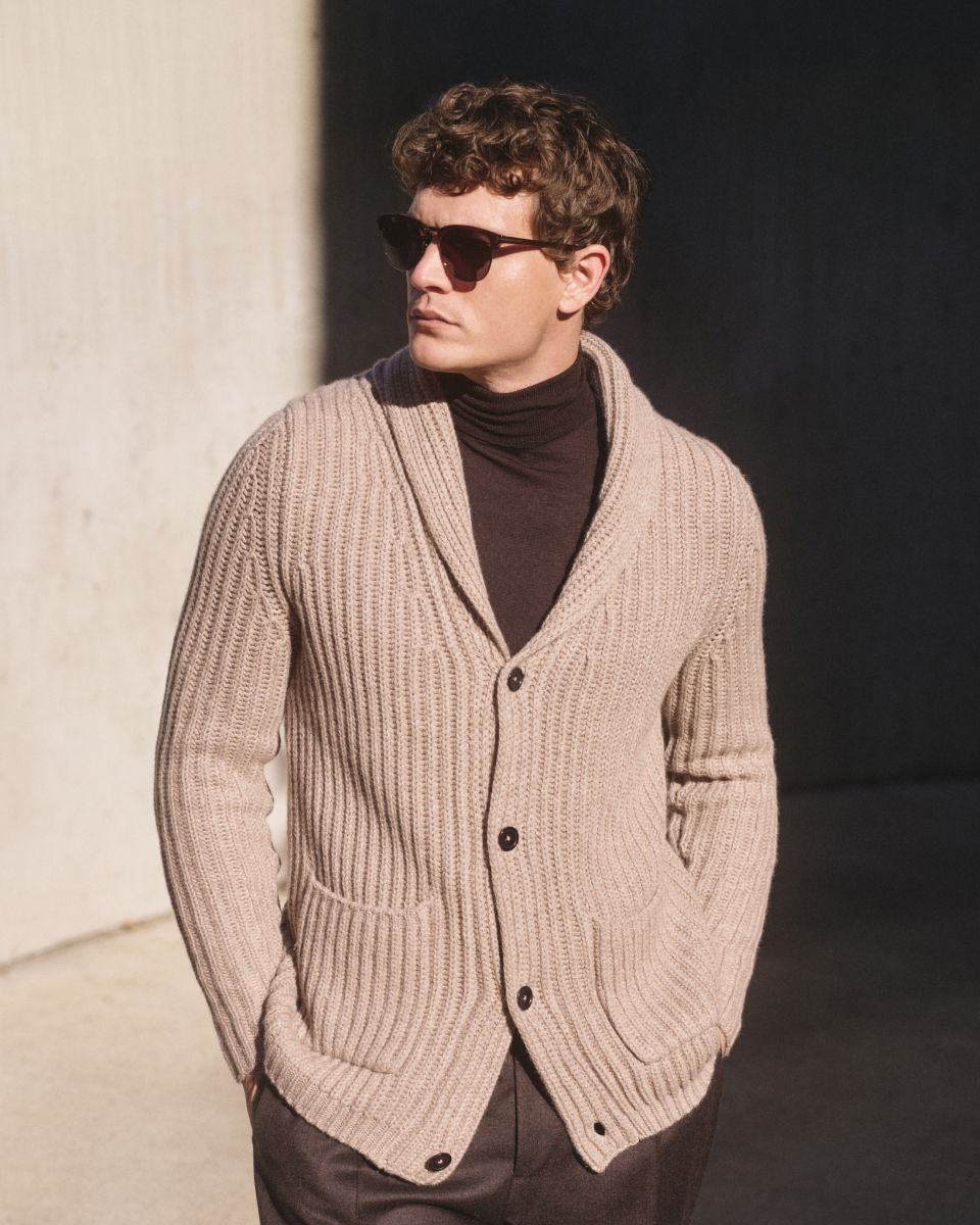 Man wearing dark sunglasses, a beige chunky knit cardigan with pockets over a dark turtleneck, standing with hands in pockets.