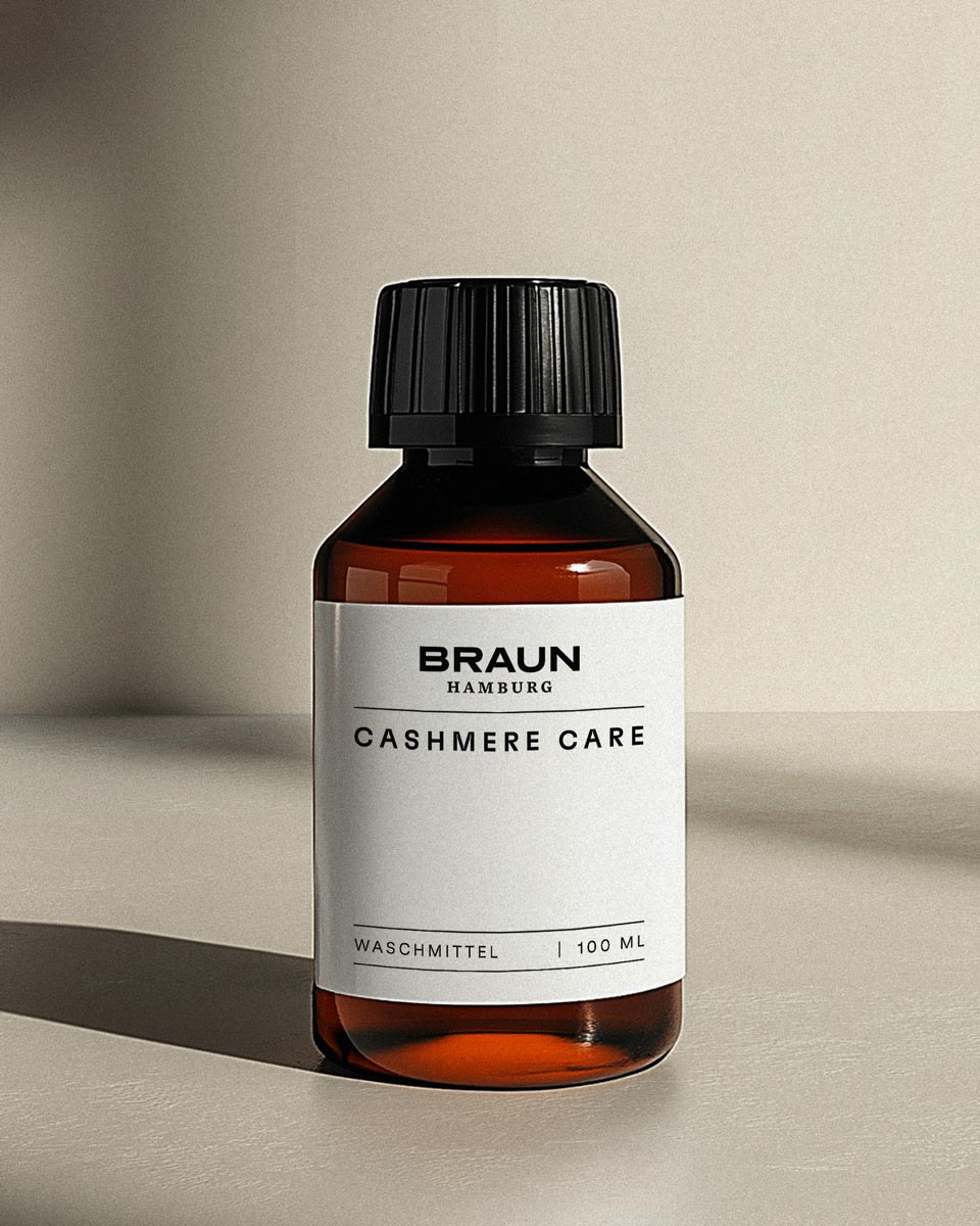 Brown translucent bottle with black cap labeled 'Braun Hamburg Cashmere Care Waschmittel 100 ml' on a beige surface.