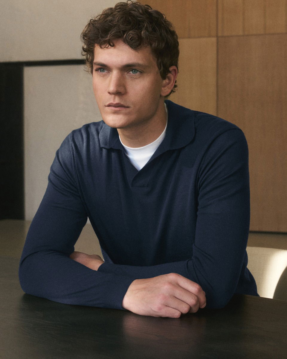 Junger Mann mit lockigem Haar sitzt am Tisch, trägt dunkelblauen Pullover über weißem T-Shirt, Blick nach rechts.