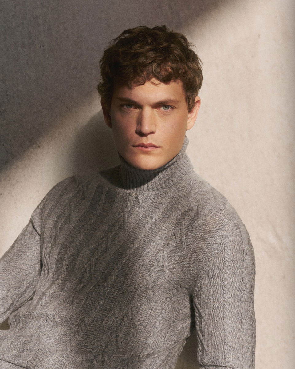 Junger Mann mit kurzen lockigen Haaren trägt hellgrauen, grob gestrickten Rollkragenpullover, neutraler Hintergrund.