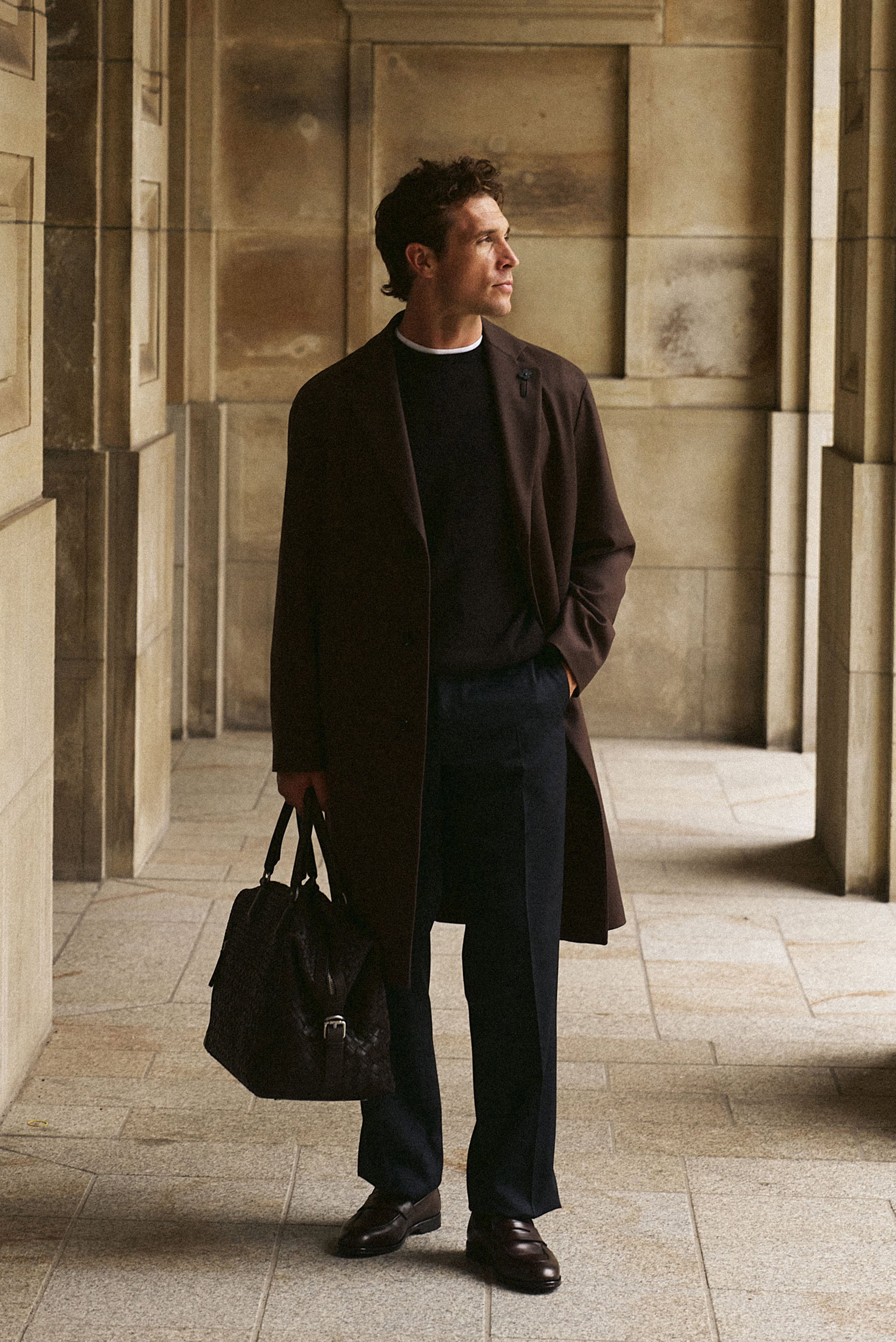 Shop by Look 01 Denis Cebulec in Lardini for BRAUN Hamburg zeigt Modell in seitlicher Nahaufnahme mit dunkelbraunem Wollmantel, Pullover, weißem T-Shirt, blauer Hose, Loafern und Weekender.