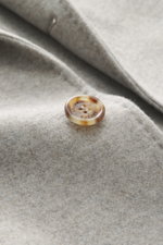 Close-up of a tortoiseshell button on the soft, grey melange fabric of the Brunello Cucinelli Cashmere Coat, shot from a top-down perspective. Elegant understatement in luxurious material quality: The super soft cashmere coat from BRUNELLO CUCINELLI becom