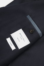 Close-up top-down view of navy Boglioli Suit 'K. Jacket' fabric showing fine tonal herringbone pattern, button, and labels.