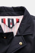 Nahaufnahme der Thom Browne Daunenjacke navy von oben, zeigt glatten Microfaserstoff, fallendes Revers, goldene Knöpfe und Tricolore-Ripsband.