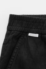 Close-up top view of 04651/ A trip in a bag Leinen-Jogpants 'Linen Pants' schwarz showing elastic waistband, French pocket, pure linen fabric with typical texture, tapered fit, regular thigh width, ankle length, straight cut, small woven label, and patch 