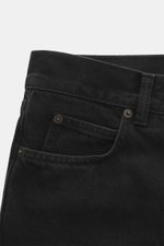 Close-up front left side view of The Row Jeans 'Vanya' black in textured black denim with tonal fading, soft touch, tapered fit, regular thigh, ankle length, straight cut, zip fly, and five-pocket style.