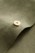 Nahaufnahme eines cremefarbenen Knopfes auf olivgrünem Leinenstoff, Detailaufnahme des von Braun Leinenhemd Haifisch-Kragen oliv, zeigt die strukturierte, luftige Leinwandbindung und weichen Griff.