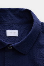 Close-up front view of the navy 04651/ A trip in a bag Seersucker shirt Kent collar navy, showing textured seersucker cotton fabric, Kent collar, single button, and brand label inside collar. Effortlessly casual: This shirt made of pure cotton by 04651/ A