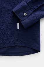 Close-up of the hem and cuff of the 04651/ A trip in a bag Seersucker shirt Kent collar navy, showing textured navy seersucker fabric, buttoned barrel cuff, side seam label, and short hem vents, photographed from above.

Description: <p><span class="_fade