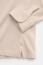 Nahaufnahme des Ärmels und Saums vom 04651/ A trip in a bag Piqué-Hemd schmaler Kragen beige aus Baumwoll-Piqué, garment dyed, mit französischer Knopfleiste und kleinem Weblabel, fotografiert aus schräger Vogelperspektive.