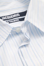 Close-up photo from above of the Jacquemus Casual shirt kent collar light blue/white striped showing pure cotton fabric, soft smooth feel, fine stripe pattern with logo woven detail, oversized fit, Kent collar, concealed button placket, initial patch on t