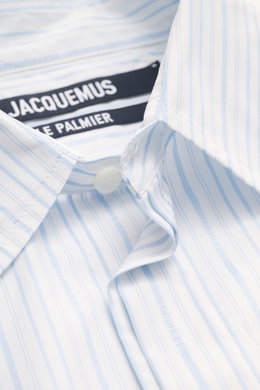 Close-up photo from above of the Jacquemus Casual shirt kent collar light blue/white striped showing pure cotton fabric, soft smooth feel, fine stripe pattern with logo woven detail, oversized fit, Kent collar, concealed button placket, initial patch on t