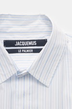 Close-up front view of Jacquemus Casual shirt kent collar light blue/white striped, showing pure cotton fabric, soft smooth feel, fine stripe pattern with logo woven detail, Kent collar, concealed button placket, initial patch on chest, pin tuck, classic 