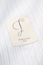 Jacquemus Casual shirt kent collar light blue/white striped shown in close-up from above, featuring pure cotton, soft smooth feel, fine stripe pattern with woven logo detail, oversized fit, Kent collar, concealed button placket, initial patch on chest, pi