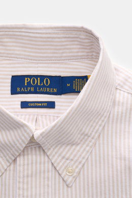 Nahaufnahme des Polo Ralph Lauren Oxford-Hemd Button-Down-Kragen beige/weiß gestreift, sportliches Baumwollhemd mit feinem Streifenmuster, Button-Down-Kragen und Logo-Stickerei, fotografiert von oben frontal.