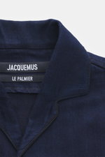 Nahaufnahme des offenen Kragens des Jacquemus Denim Kurzarmhemd dunkelblau aus reine Baumwolle, Regular Fit mit aufgesetzter Brusttasche und dekorativer J-Schlaufe, fotografiert aus schräger Frontperspektive.