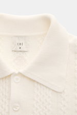 Nahaufnahme des Ché Kurzarm-Strickpolo 'Santino' creme von oben, zeigt grobes, asymmetrisches Strickmuster, Polokragen, Drei-Knopf-Leiste, Baumwolle, Regular Fit, flexible Rippbündchen und tonales CHÉ-Weblabel.