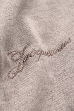 Close-up image of the Jacquemus Merino Knit Polo 'Arlequin' taupe/grey brown melange showing slightly elastic merino wool fabric with logo stitching on the hem, photographed from an angled top view. Features include regular fit, color-contrasted polo coll