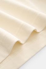 Close-up image from a top angle showing the fabric texture of the Jacquemus Crew Neck T-Shirt cream, featuring pure cotton, soft touch, slim fit, and grosgrain with logo stitching on the chest.