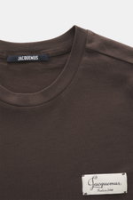 Close-up front view of Jacquemus Crew neck T-shirt 'Le Montaigne MC' dark brown, jersey made of pure cotton, soft touch, fine diagonal texture, slim fit, crew neckline, woven label on chest, side hem slits, extended back.