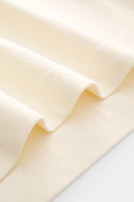 Close-up view of folded Jacquemus Crew Neck T-Shirt cream fabric showing pure cotton material, soft touch, slim fit, and grosgrain with logo stitching and print on the chest.