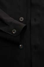 Close-up front view of the 04651/ A trip in a bag Seersucker-Overshirt schwarz, showing black seersucker fabric with garment dyed texture, slim fit design, Kent collar, five-button placket, cuff with button, classic chest pocket, and detailed stitching.