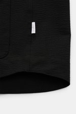 Close-up of the hem and side seam of the 04651/ A trip in a bag Seersucker-Overshirt schwarz, showing black wool and microfiber fabric texture, a small white label with "04651/" on the side seam, garment dyed, photographed from a top-down perspective. <ar