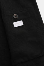 Close-up image of the 04651/ A trip in a bag Seersucker-Overshirt schwarz pocket, showing black seersucker fabric with garment-dyed texture, a single dark button, and a white label with the brand name, photographed from a top-down perspective highlighting