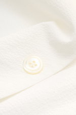Nahaufnahme eines weißen Knopfs auf dem 04651/ A trip in a bag Overshirt 'Technical Seersucker' offwhite mit strukturierter Microfaser, Slim Fit, fünffacher Knopf-Leiste, Umlegekragen und paspelierten Fronttaschen, fotografiert aus schräger Perspektive.