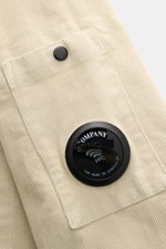 C.P. Company Cord-Overshirt sand, Nahaufnahme der Brusttasche mit Knopf, samtiger Cord, Slim Fit, Linsen-Detail seitlich, von schräg oben.