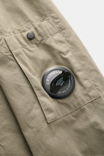 Nahaufnahme des C.P. Company Overshirt graugrün aus samtiger Baumwollgabardine mit Druckknopftasche und labeltypischem Linsen-Detail.