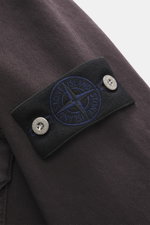 Close-up view of the Stone Island Jeansjacke 'Light Bull Denim-TC' dunkelbraun sleeve, showing the black compass badge with blue embroidery and silver buttons, highlighting the robust pure cotton denim fabric with a slightly textured, garment-dyed finish.