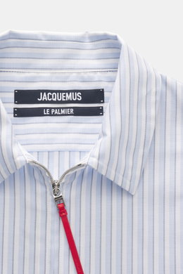 Close-up front view of Jacquemus Light blue/white striped overshirt showing turn-down collar, two-way zipper with red pull, decorative J-hook, subtle logo stitching, classic sport cuff, oversized fit, slightly textured pure cotton oxford fabric.