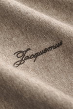 Close-up view of Jacquemus Overshirt taupe meliert fabric showing logo-stitching, made of reine Schurwolle with weicher Griff, Oversized Fit, Umlegekragen, Zwei-Wege-Reißverschluss, decorative Leder-Zipperanhänger, klassische Sportmanschette, and gerader 