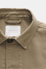 Nahaufnahme des Kragens und Etiketts von 04651/ A trip in a bag Overshirt 'Workwear Jacket' taupe, reine Baumwolle, Slim Fit, Unconstructed Design, Fünf-Knopf-Leiste, Umlegekragen, leicht griffige Haptik, fotografiert aus frontaler Perspektive.
