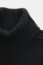 Nahaufnahme aus schräger Perspektive des Belstaff Rollkragenpullover 'Watch' schwarz mit grobem Strickbild, reiner Wolle, weichem Griff, Slim Fit, Rollkragen, Rippstrick-Elementen, flexible Rippbündchen und BELSTAFF-Weblabel auf der Brust.