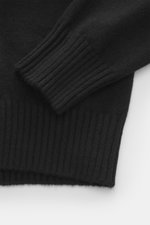 Nahaufnahme des schwarzen Ami Paris Cashmere Rollkragenpullovers mit weichem Griff, mittelfeinem Strickbild, Regular Fit, anliegendem Rollkragen, Logo-Stitching am Saum und komfortablen Rippbündchen, perspektivisch von der Seite fotografiert.