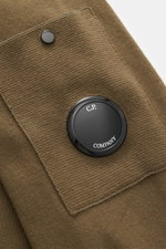 Nahaufnahme eines olivgrünen C.P. Company Rundhalspullovers, fotografiert schräg von oben, mit Fokus auf Tasche und rundem Linsen-Detail.