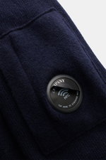 C.P. Company Troyer navy, Nahaufnahme von weichem Strick mit labeltypischem Linsen-Detail und Zipper-Tasche am Oberarm, perspektivisch schräg.